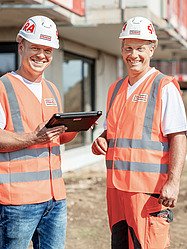 Mitarbeiter auf der Baustelle Foto von drei Männern auf der Baustelle, die vor Baugeräten stehen und Arbeitskleidung anhaben