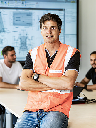 Einstiegsmöglichkeiten bei STRABAG Mitarbeiter:innen sitzen im Hintergrund während ein Mitarbeiter im Vordergrund in die Kamera lächelt